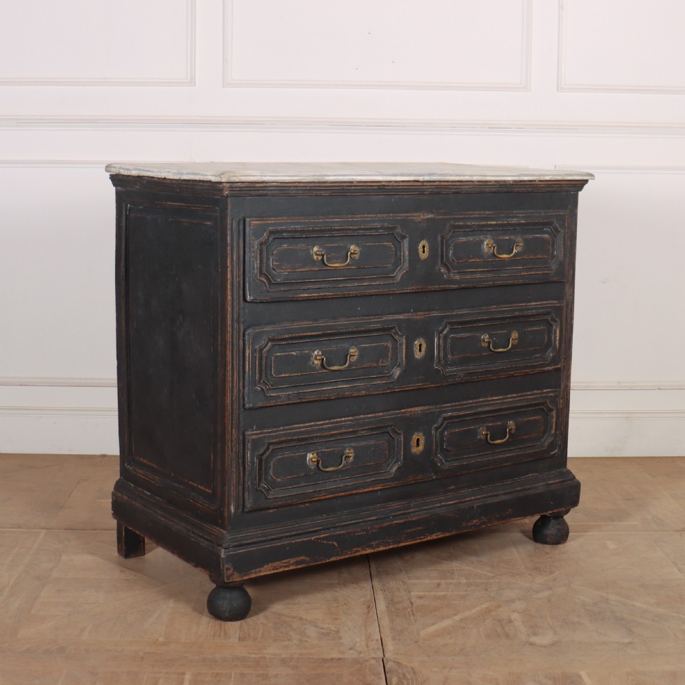 An 18th-century French painted black commode featuring three long drawers with moulded panels, brass drop handles, and a grey marble top.