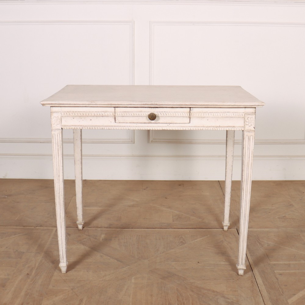 An off-white painted Swedish Gustavian side table featuring slim fluted legs, carved corner rosettes, and a single frieze drawer with a brass pull knob.