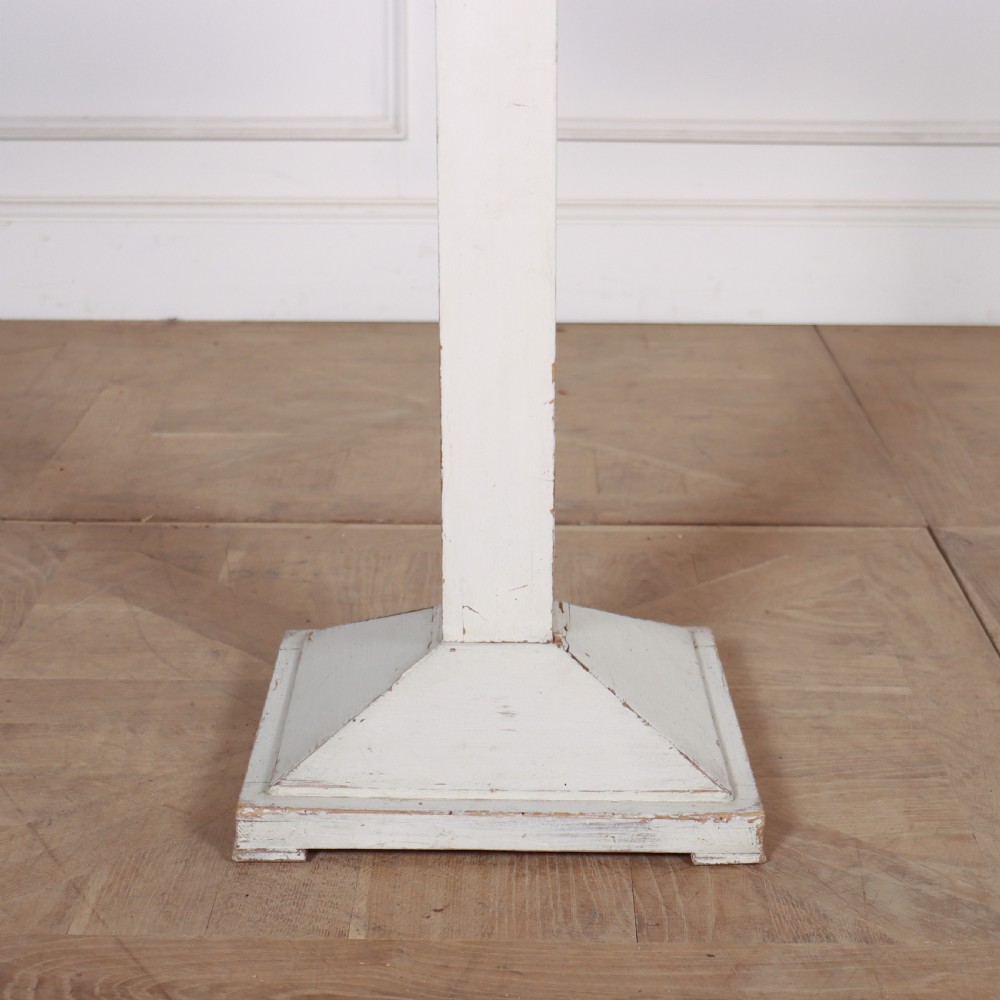 An off-white painted Swedish pedestal table featuring a circular top, a square central column, and a stepped plinth base with visible paint wear along the edges. View V