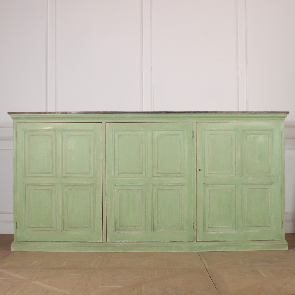 A low sage green painted wooden linen cupboard with three panelled doors and a dark wood top.