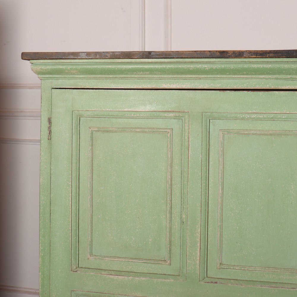 A low sage green painted wooden linen cupboard with three panelled doors and a dark wood top. View II