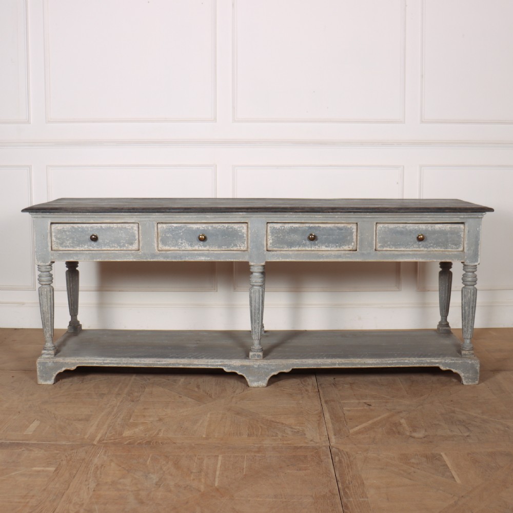 A long French style drapers console table featuring a distressed grey painted frame, dark wooden top, four drawers, fluted turned legs, and a low pot board shelf.