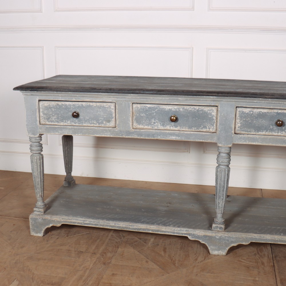 A long French style drapers console table featuring a distressed grey painted frame, dark wooden top, four drawers, fluted turned legs, and a low pot board shelf. View II