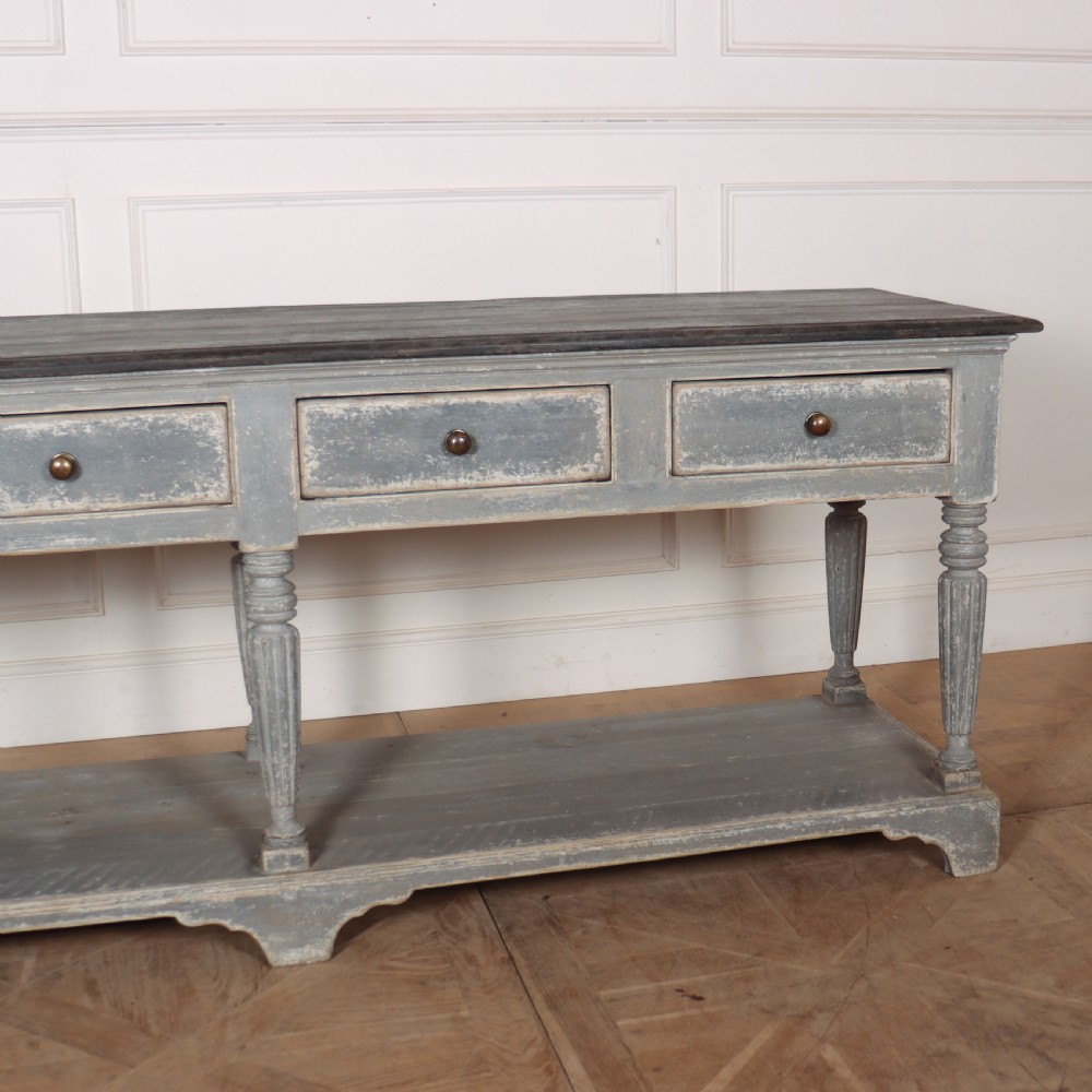 A long French style drapers console table featuring a distressed grey painted frame, dark wooden top, four drawers, fluted turned legs, and a low pot board shelf. View III