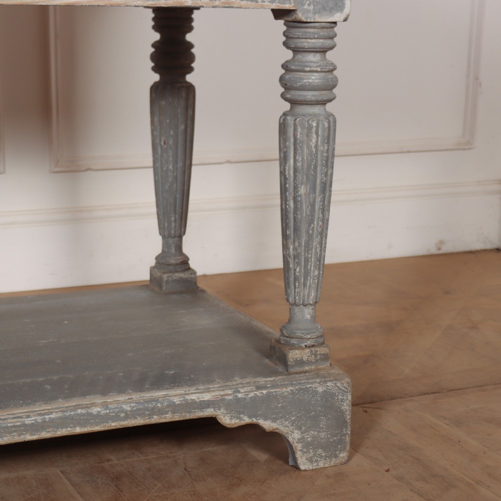 A long French style drapers console table featuring a distressed grey painted frame, dark wooden top, four drawers, fluted turned legs, and a low pot board shelf. View IV