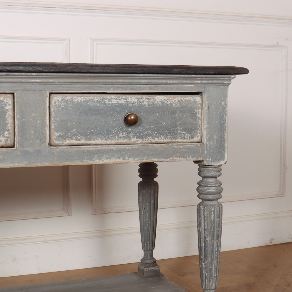 A long French style drapers console table featuring a distressed grey painted frame, dark wooden top, four drawers, fluted turned legs, and a low pot board shelf. View V