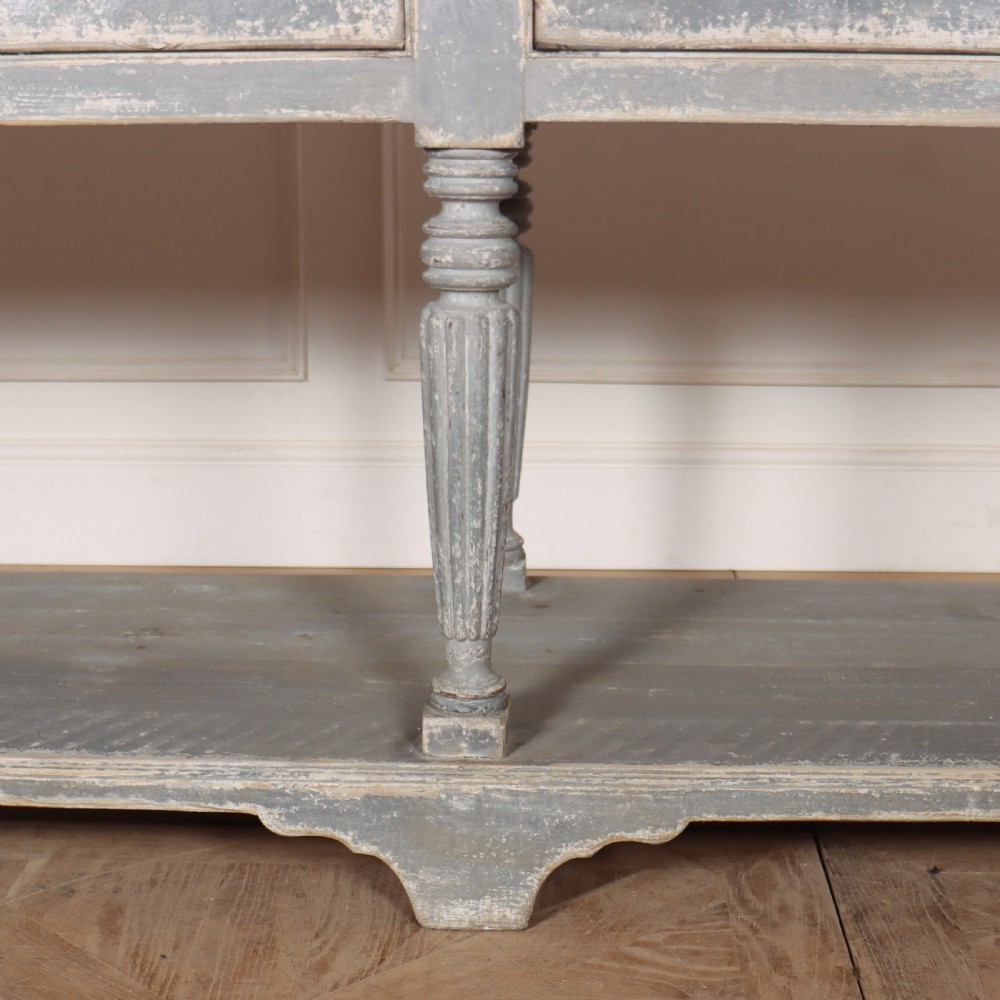 A long French style drapers console table featuring a distressed grey painted frame, dark wooden top, four drawers, fluted turned legs, and a low pot board shelf. View VI