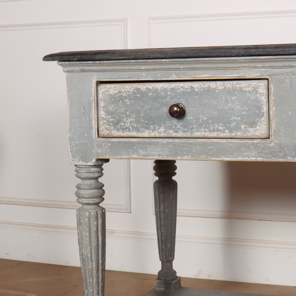 A long French style drapers console table featuring a distressed grey painted frame, dark wooden top, four drawers, fluted turned legs, and a low pot board shelf. View VII