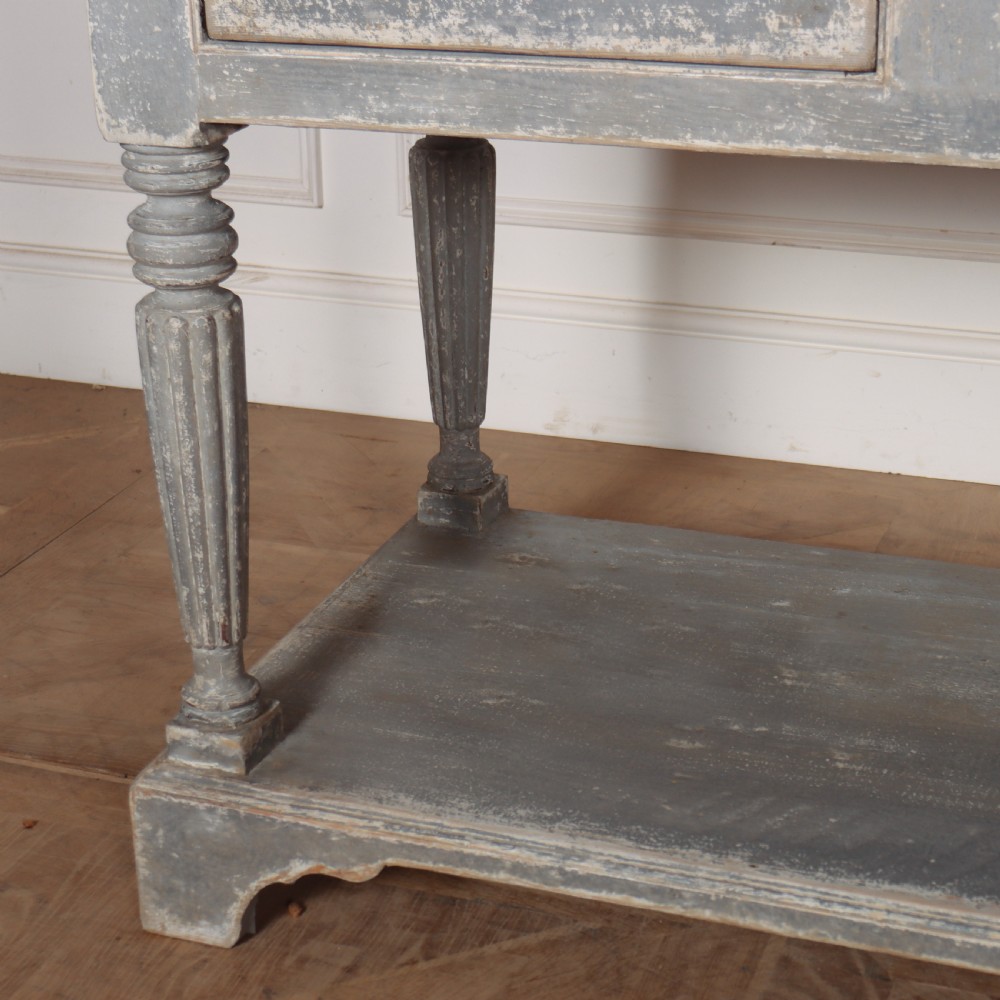 A long French style drapers console table featuring a distressed grey painted frame, dark wooden top, four drawers, fluted turned legs, and a low pot board shelf. View VIII