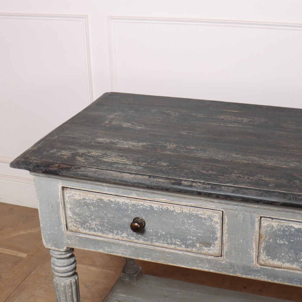 A long French style drapers console table featuring a distressed grey painted frame, dark wooden top, four drawers, fluted turned legs, and a low pot board shelf. View IX