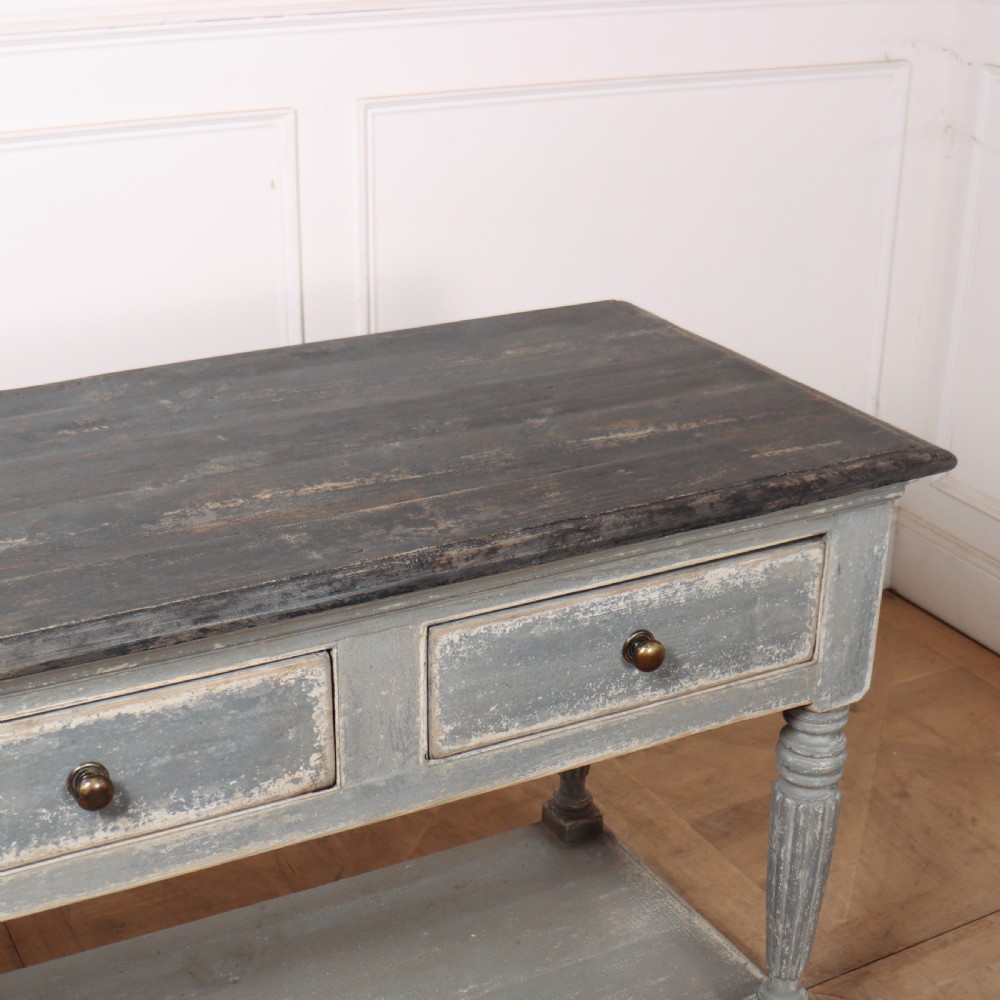 A long French style drapers console table featuring a distressed grey painted frame, dark wooden top, four drawers, fluted turned legs, and a low pot board shelf. View X