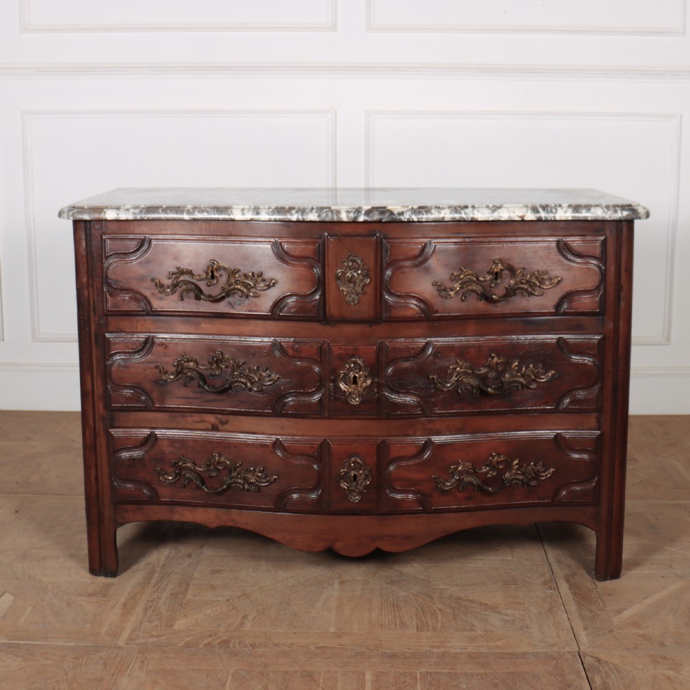 An 18th-century French serpentine commode in warm brown timber featuring three drawers with carved panels, ornate brass handles, and a grey veined marble top.