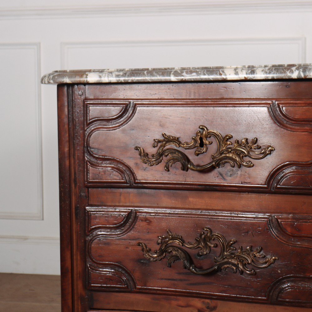 An 18th-century French serpentine commode in warm brown timber featuring three drawers with carved panels, ornate brass handles, and a grey veined marble top. View II