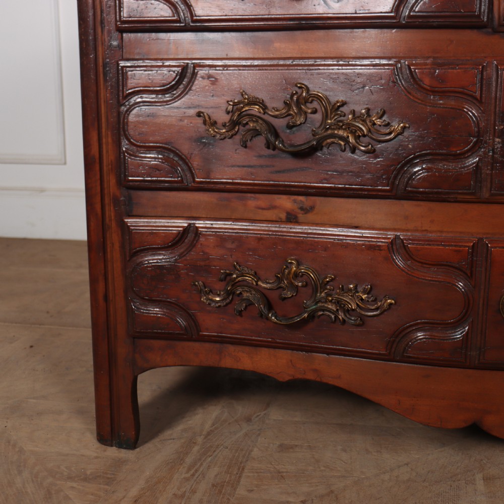 An 18th-century French serpentine commode in warm brown timber featuring three drawers with carved panels, ornate brass handles, and a grey veined marble top. View III