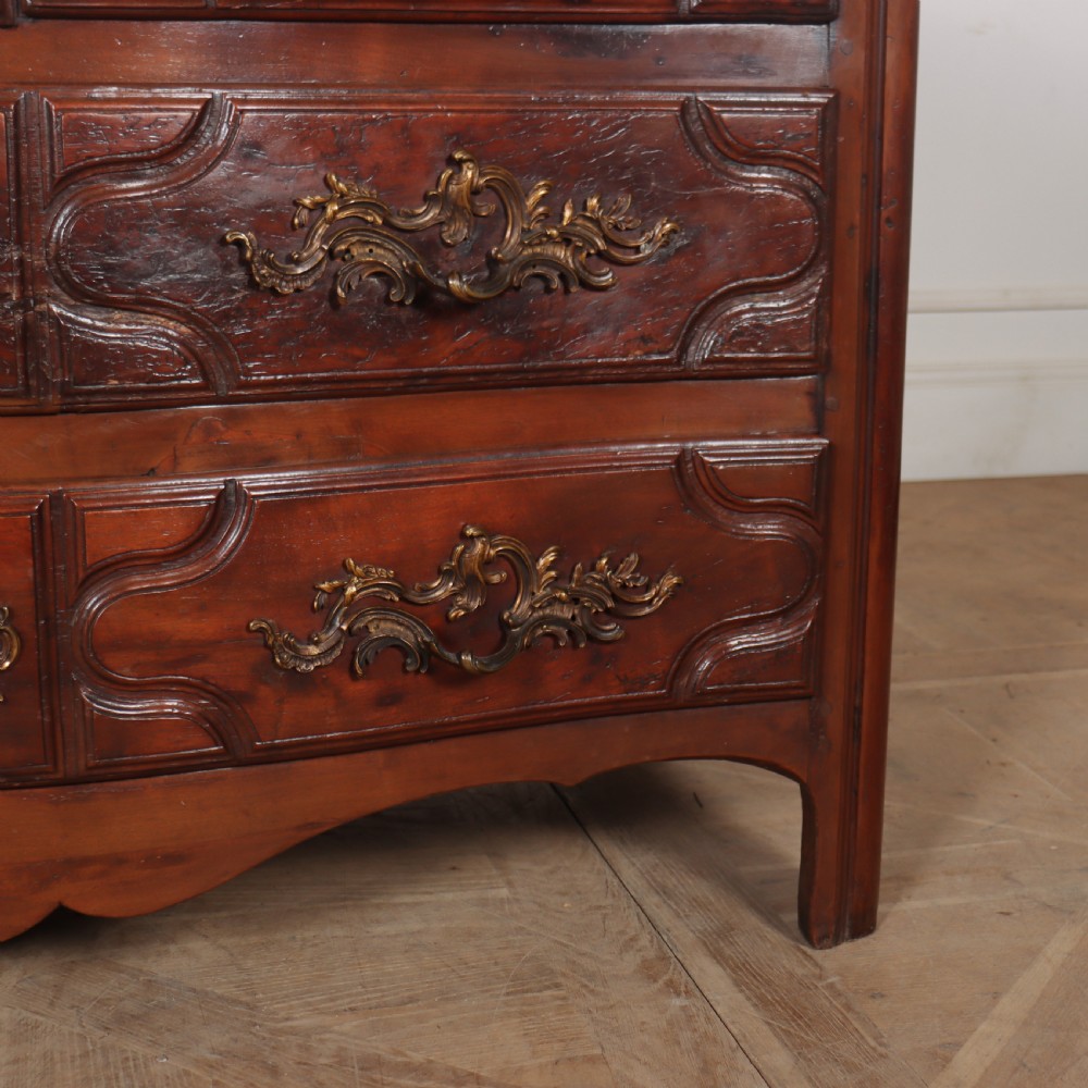An 18th-century French serpentine commode in warm brown timber featuring three drawers with carved panels, ornate brass handles, and a grey veined marble top. View IV