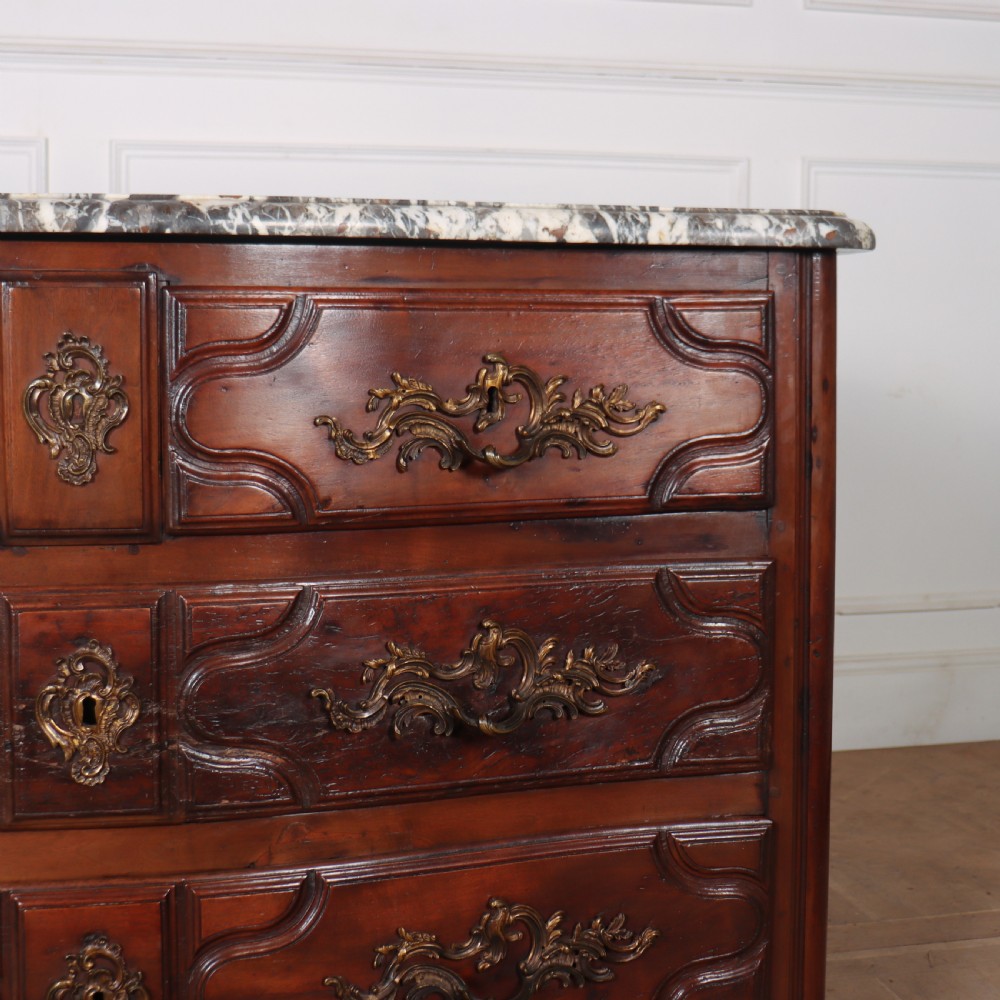 An 18th-century French serpentine commode in warm brown timber featuring three drawers with carved panels, ornate brass handles, and a grey veined marble top. View V
