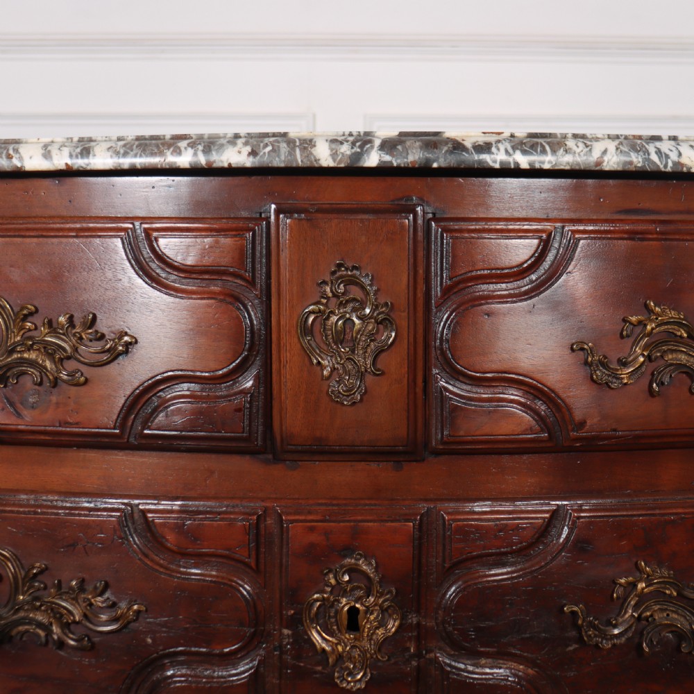An 18th-century French serpentine commode in warm brown timber featuring three drawers with carved panels, ornate brass handles, and a grey veined marble top. View VI