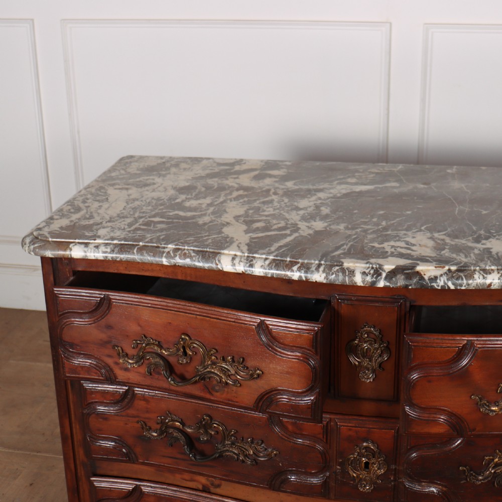 An 18th-century French serpentine commode in warm brown timber featuring three drawers with carved panels, ornate brass handles, and a grey veined marble top. View VII