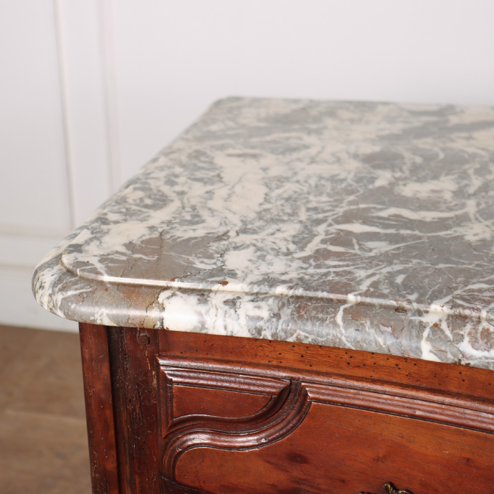 An 18th-century French serpentine commode in warm brown timber featuring three drawers with carved panels, ornate brass handles, and a grey veined marble top. View VIII