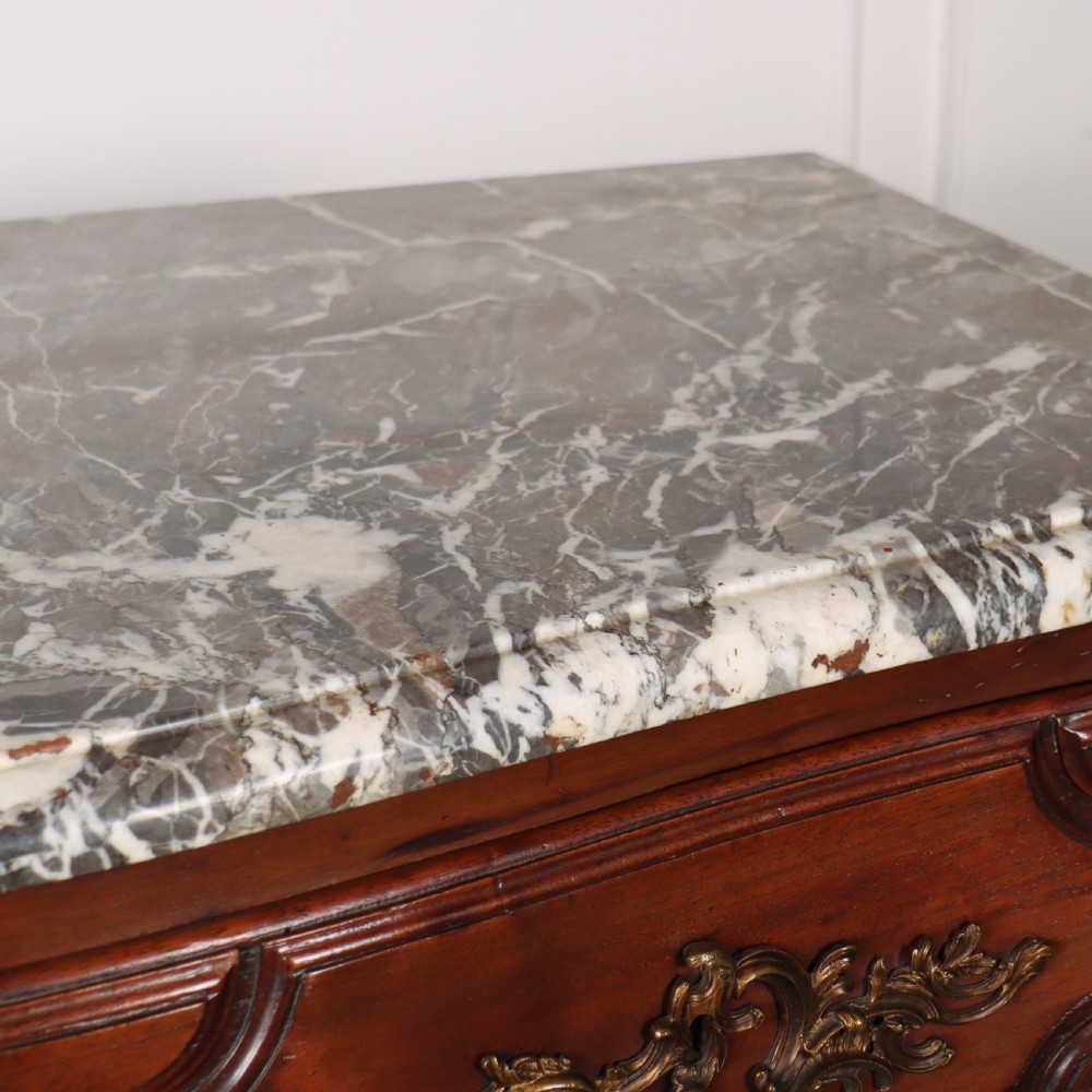 An 18th-century French serpentine commode in warm brown timber featuring three drawers with carved panels, ornate brass handles, and a grey veined marble top. View IX