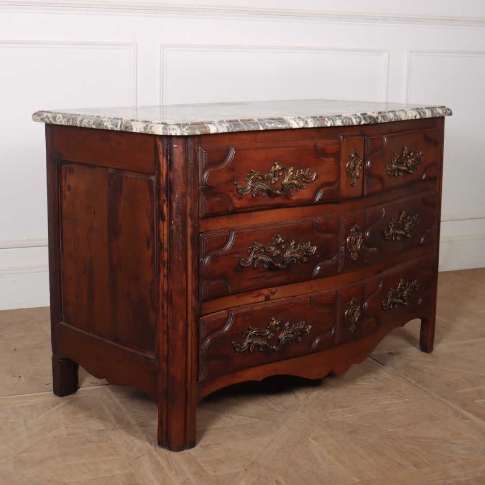 An 18th-century French serpentine commode in warm brown timber featuring three drawers with carved panels, ornate brass handles, and a grey veined marble top. View X