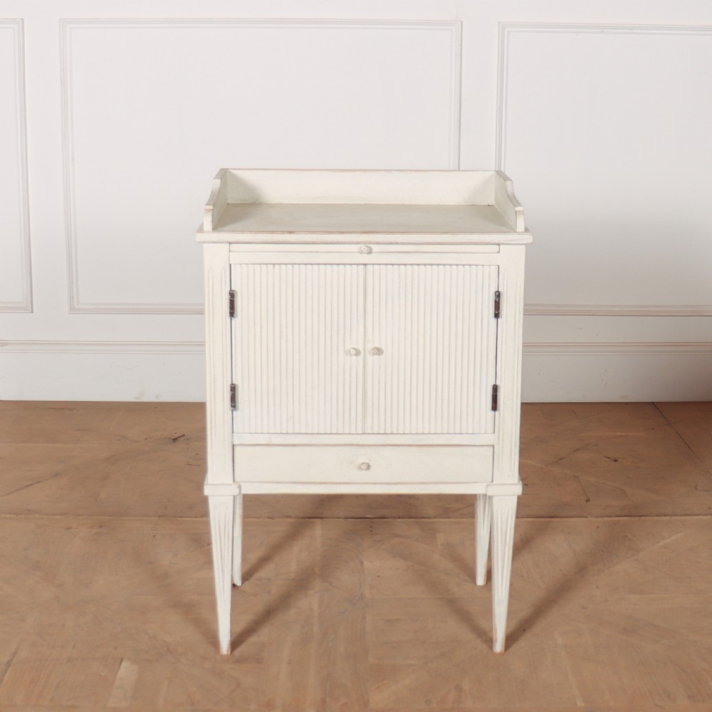 An off-white painted Gustavian style bedside cupboard featuring fluted double doors, a single lower drawer, a raised gallery top, and slender tapered legs.