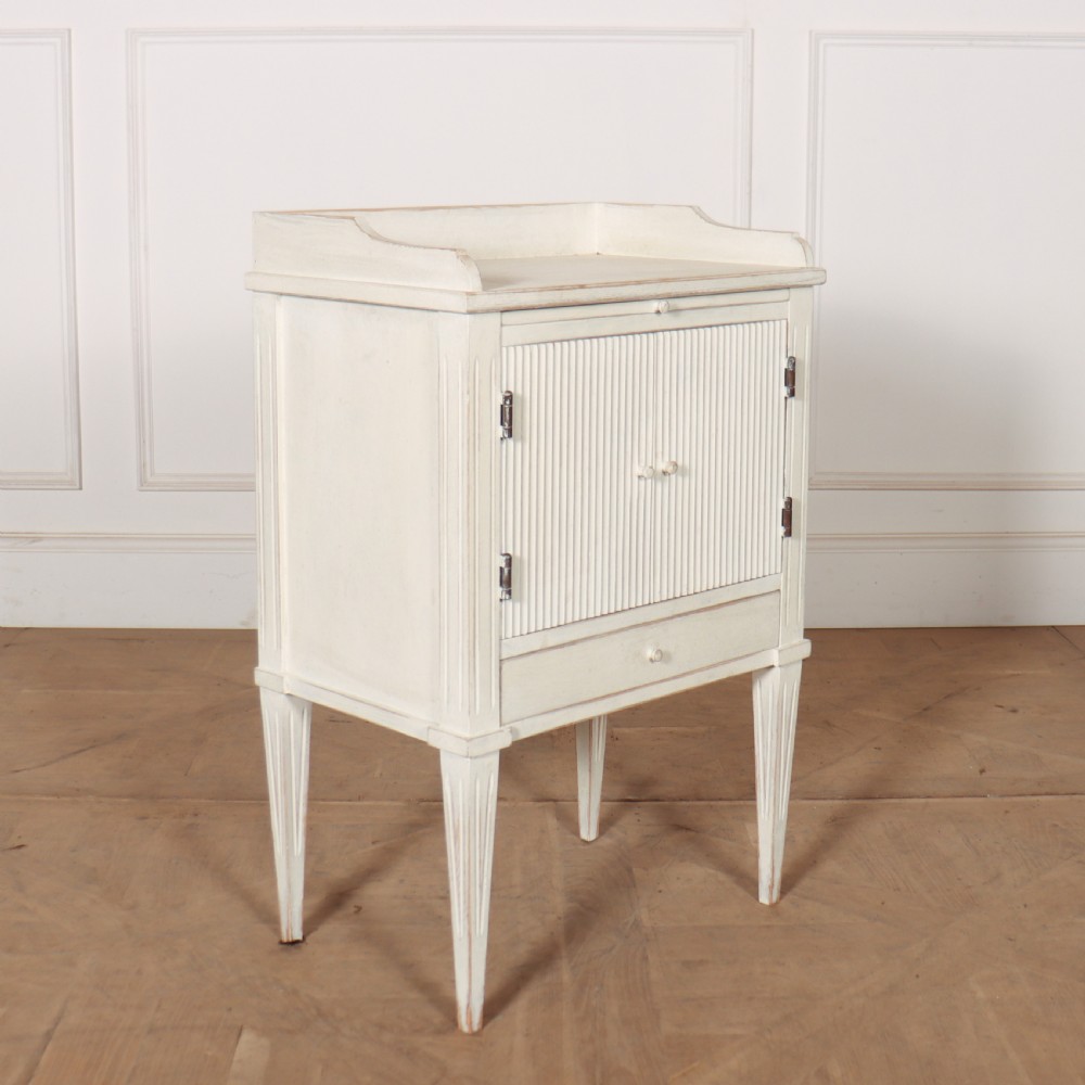 An off-white painted Gustavian style bedside cupboard featuring fluted double doors, a single lower drawer, a raised gallery top, and slender tapered legs. View II