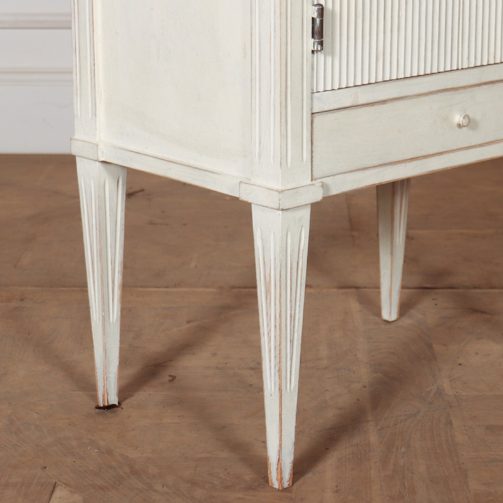 An off-white painted Gustavian style bedside cupboard featuring fluted double doors, a single lower drawer, a raised gallery top, and slender tapered legs. View III