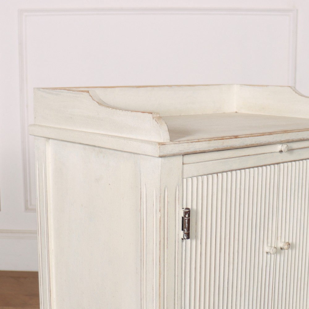 An off-white painted Gustavian style bedside cupboard featuring fluted double doors, a single lower drawer, a raised gallery top, and slender tapered legs. View IV