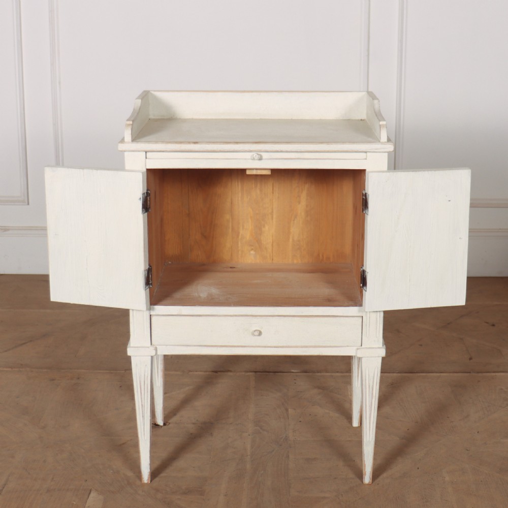 An off-white painted Gustavian style bedside cupboard featuring fluted double doors, a single lower drawer, a raised gallery top, and slender tapered legs. View V