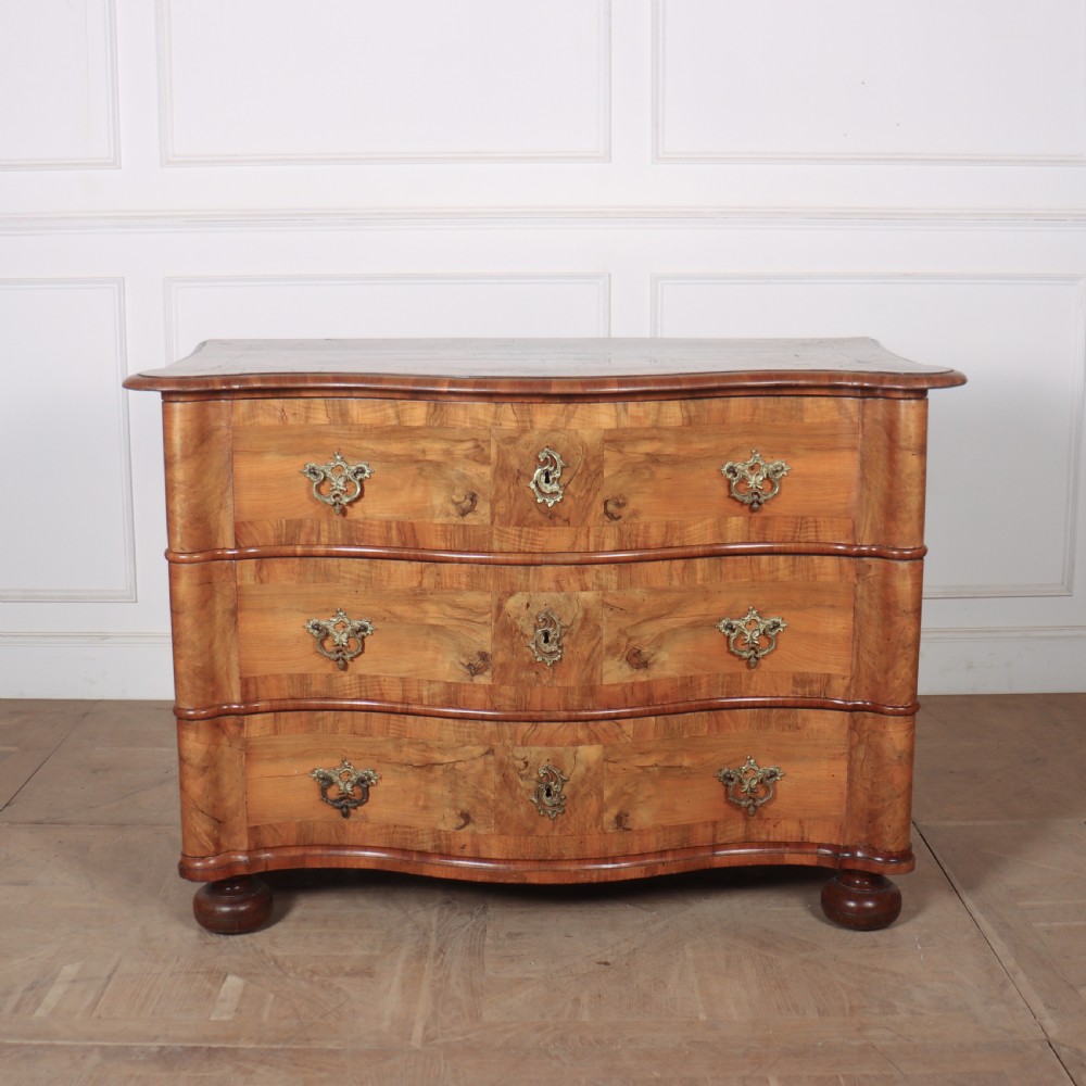 An 18th-century walnut serpentine commode featuring three long drawers with ornate brass handles and keyhole escutcheons, standing on turned bun feet.