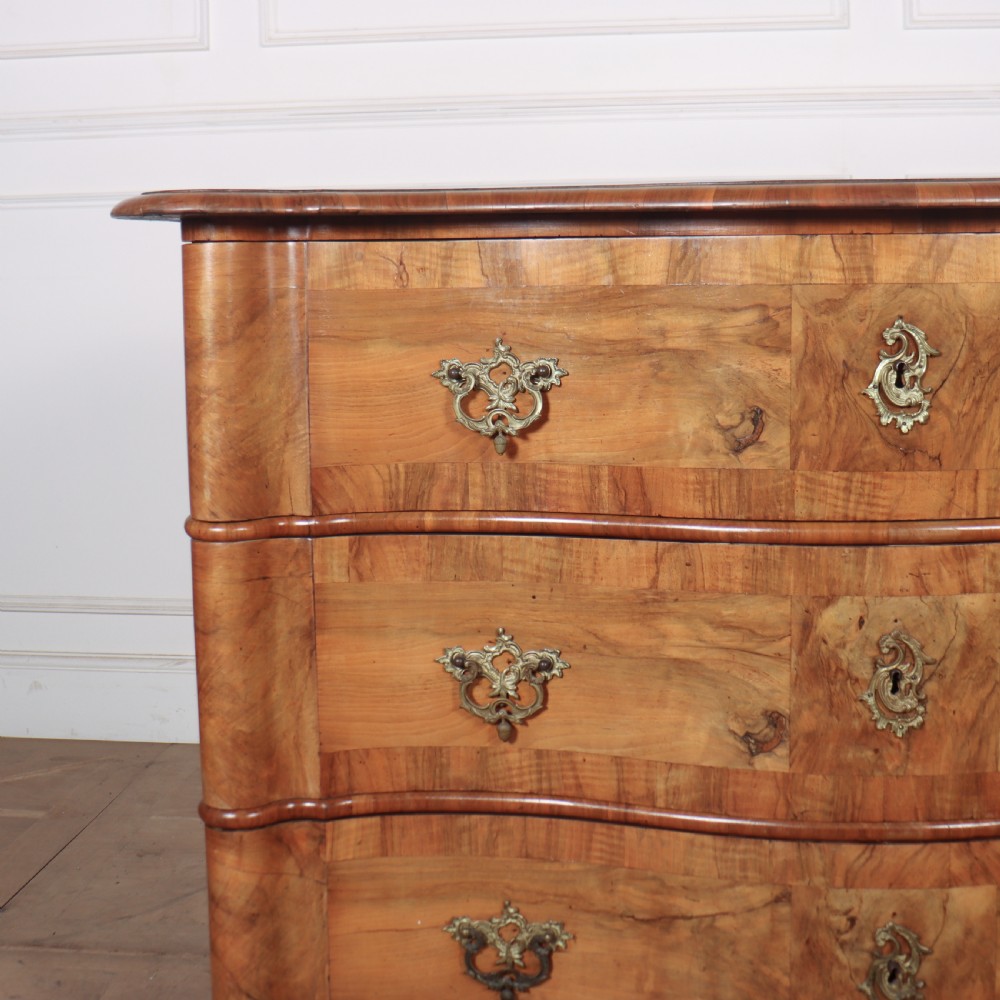 An 18th-century walnut serpentine commode featuring three long drawers with ornate brass handles and keyhole escutcheons, standing on turned bun feet. View II