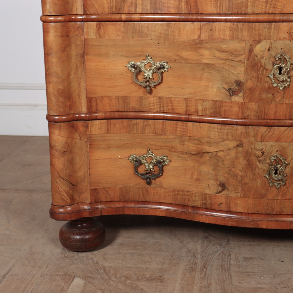 An 18th-century walnut serpentine commode featuring three long drawers with ornate brass handles and keyhole escutcheons, standing on turned bun feet. View III