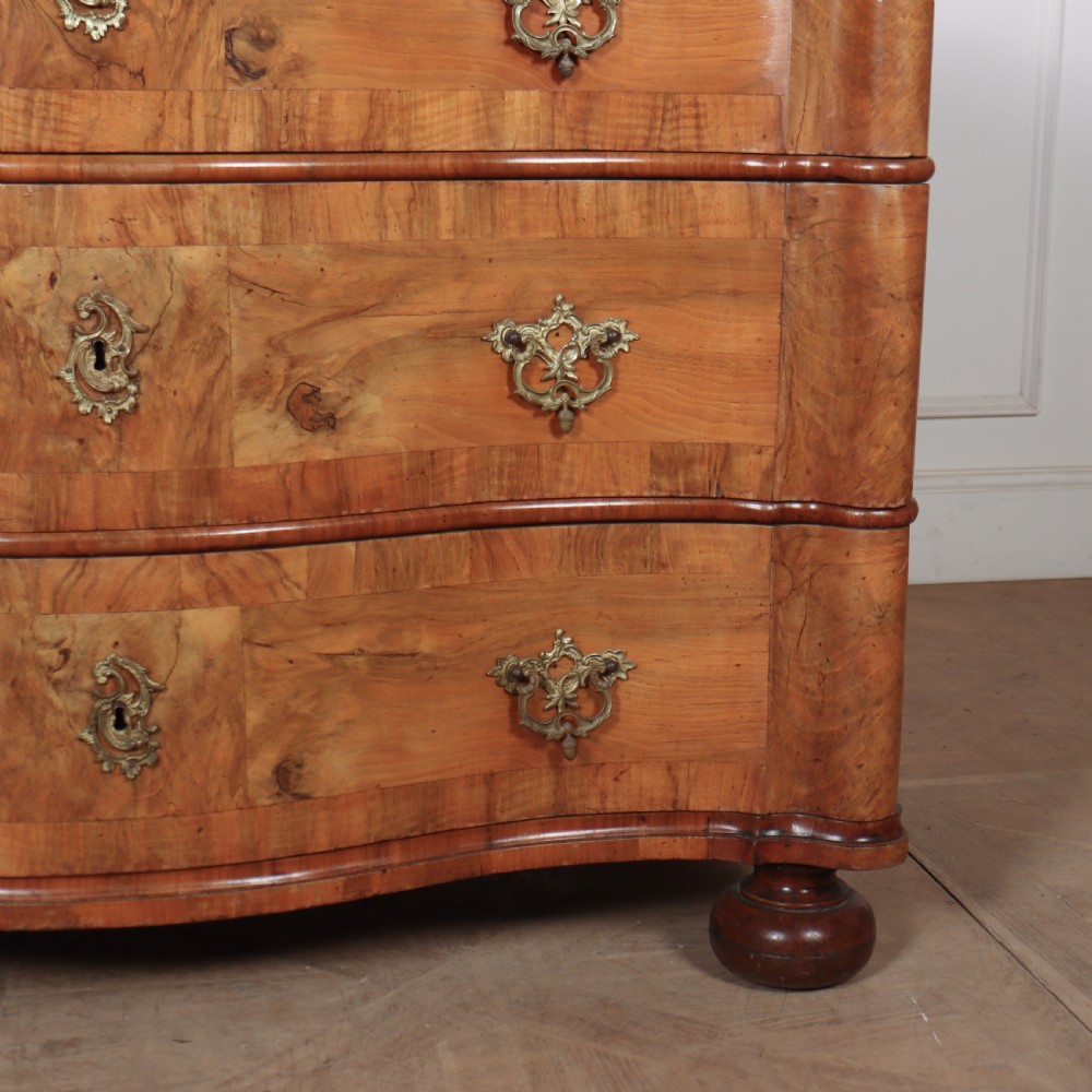 An 18th-century walnut serpentine commode featuring three long drawers with ornate brass handles and keyhole escutcheons, standing on turned bun feet. View V