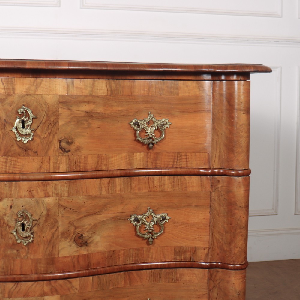 An 18th-century walnut serpentine commode featuring three long drawers with ornate brass handles and keyhole escutcheons, standing on turned bun feet. View VI
