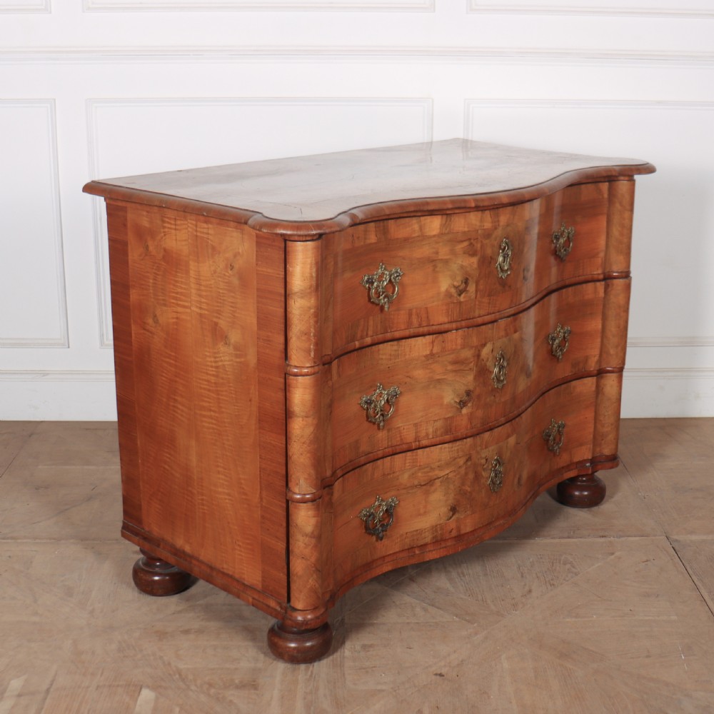 An 18th-century walnut serpentine commode featuring three long drawers with ornate brass handles and keyhole escutcheons, standing on turned bun feet. View VII