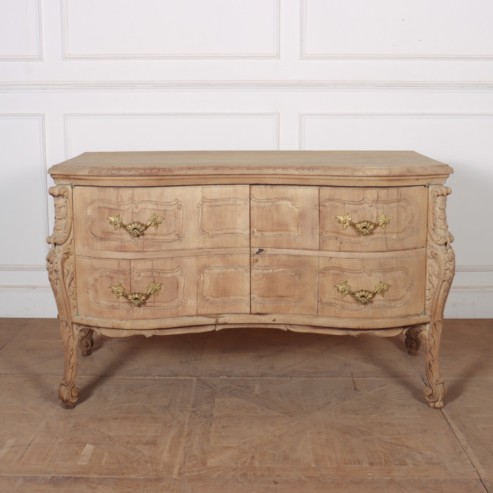 An antique French bleached oak commode featuring a serpentine front, carved rocaille mouldings, drawers with brass pulls, and cabriole feet.