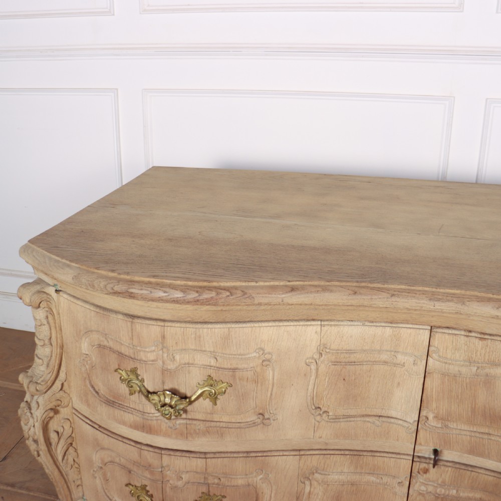 An antique French bleached oak commode featuring a serpentine front, carved rocaille mouldings, drawers with brass pulls, and cabriole feet. View VI