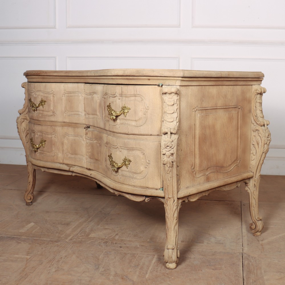An antique French bleached oak commode featuring a serpentine front, carved rocaille mouldings, drawers with brass pulls, and cabriole feet. View VII