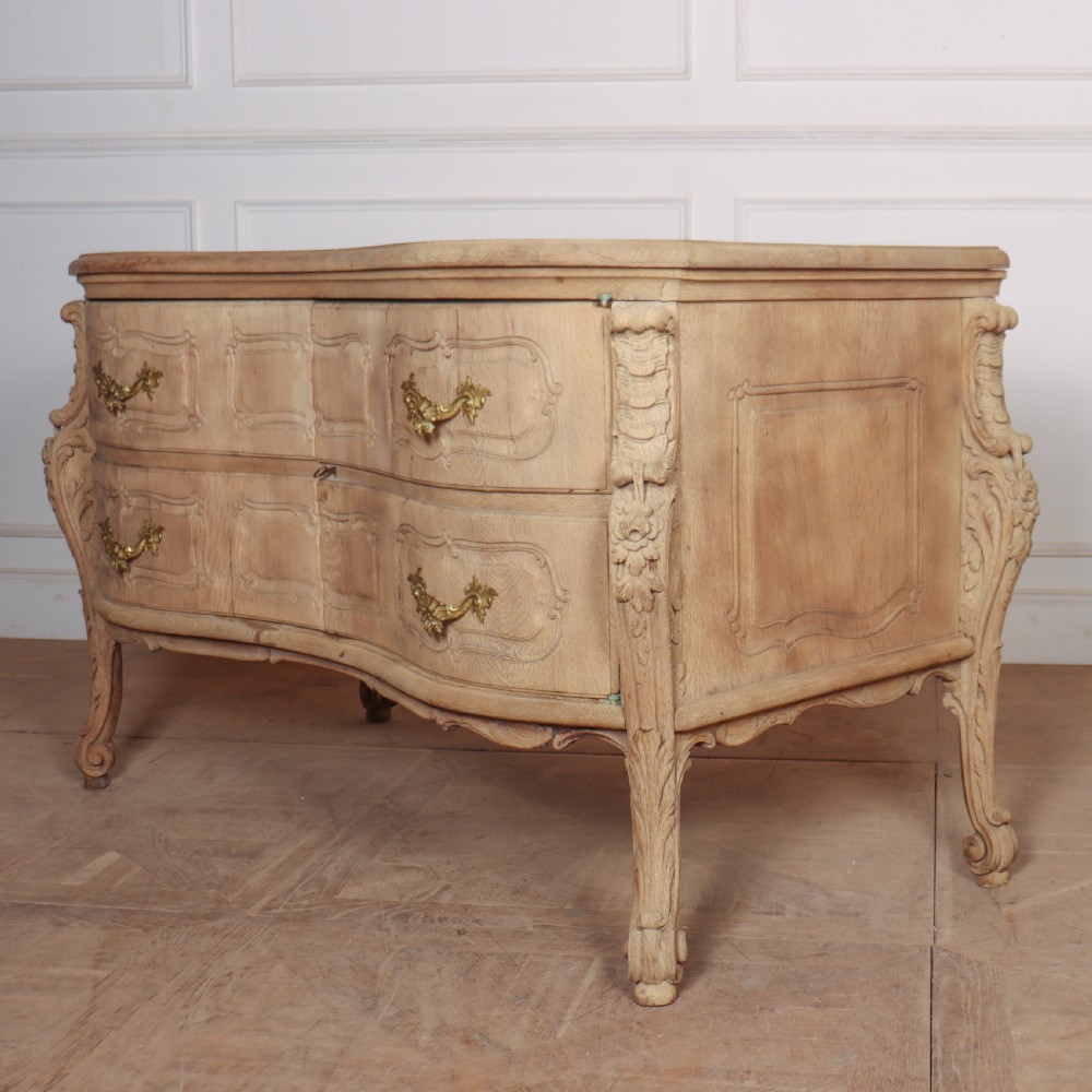 An antique French bleached oak commode featuring a serpentine front, carved rocaille mouldings, drawers with brass pulls, and cabriole feet. View VIII