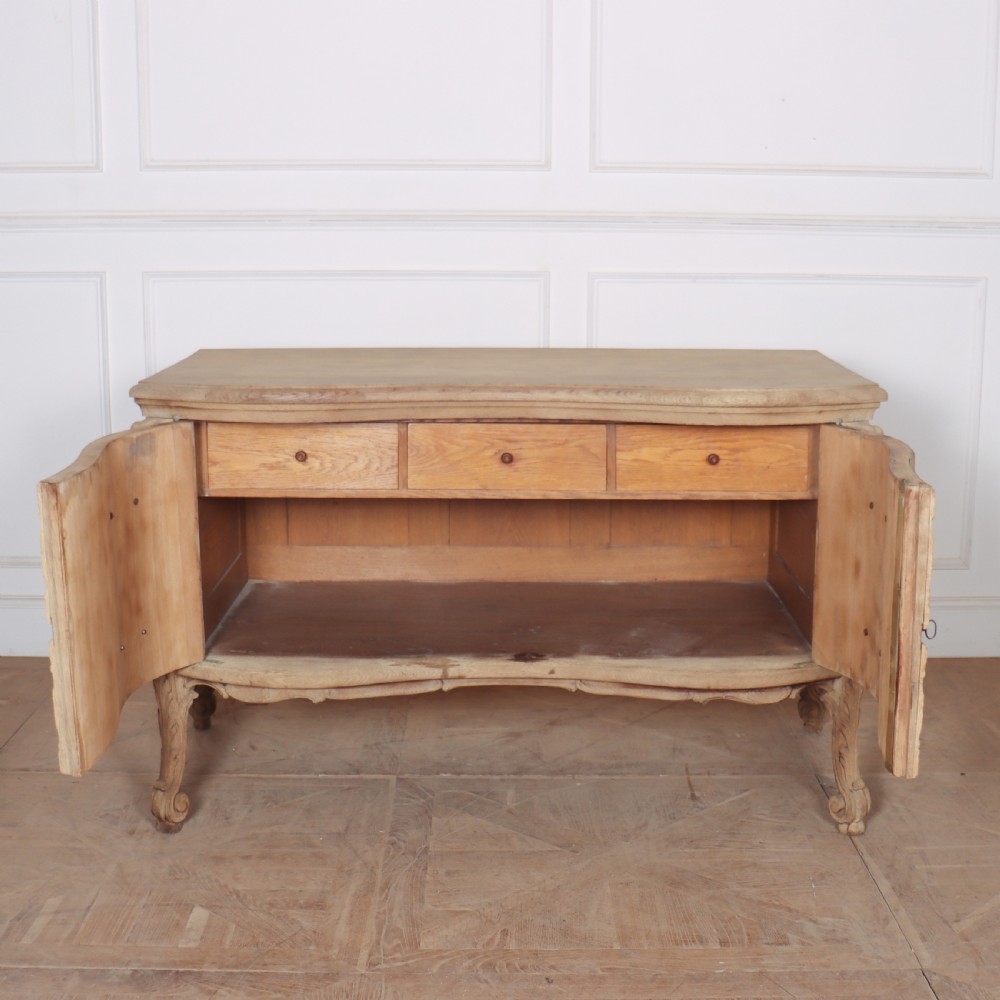 An antique French bleached oak commode featuring a serpentine front, carved rocaille mouldings, drawers with brass pulls, and cabriole feet. View X