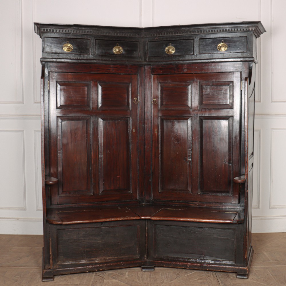 A dark stained oak corner bacon settle featuring high panelled cupboard doors, upper drawers with brass handles, and an integrated bench seat.
