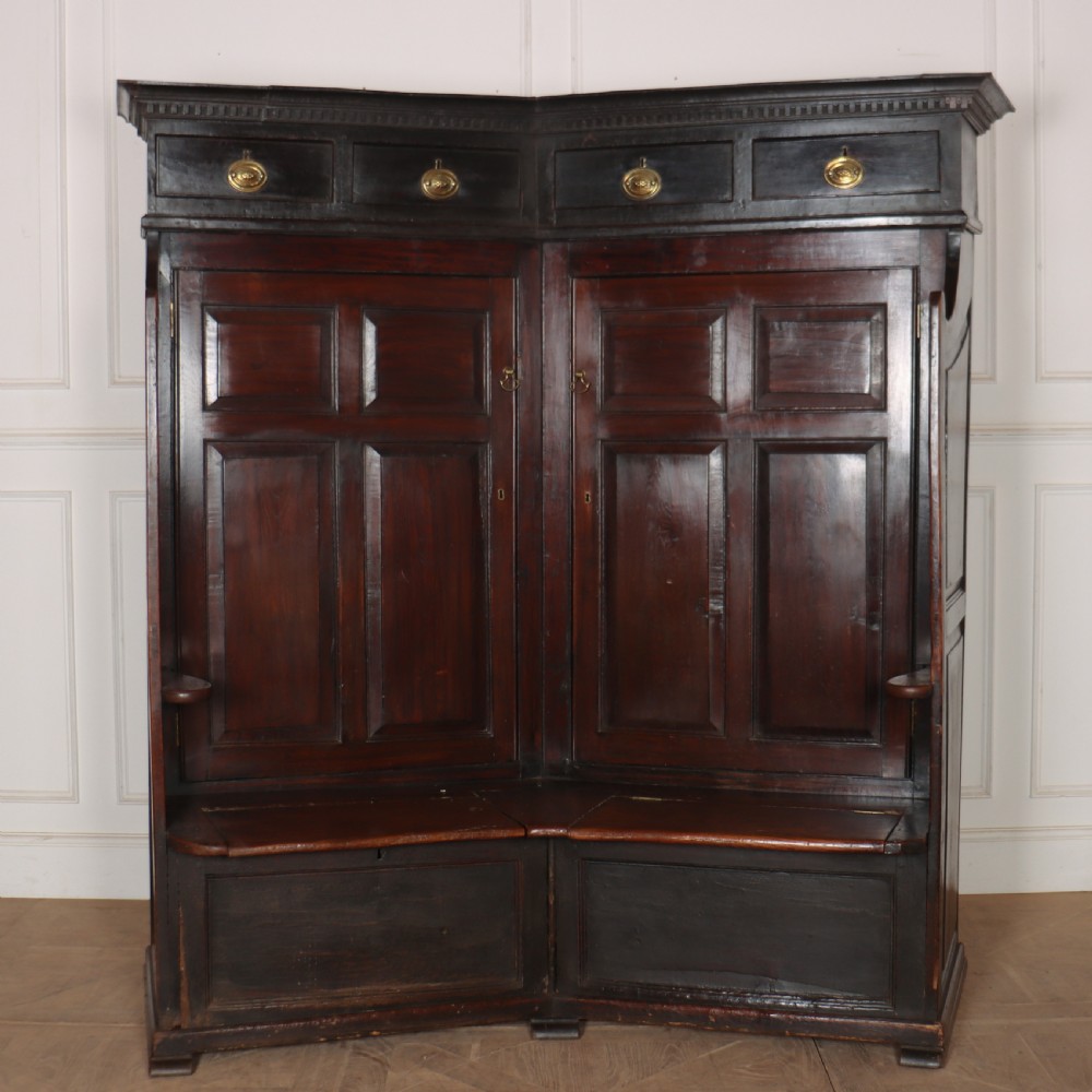 A dark stained oak corner bacon settle featuring high panelled cupboard doors, upper drawers with brass handles, and an integrated bench seat. View II