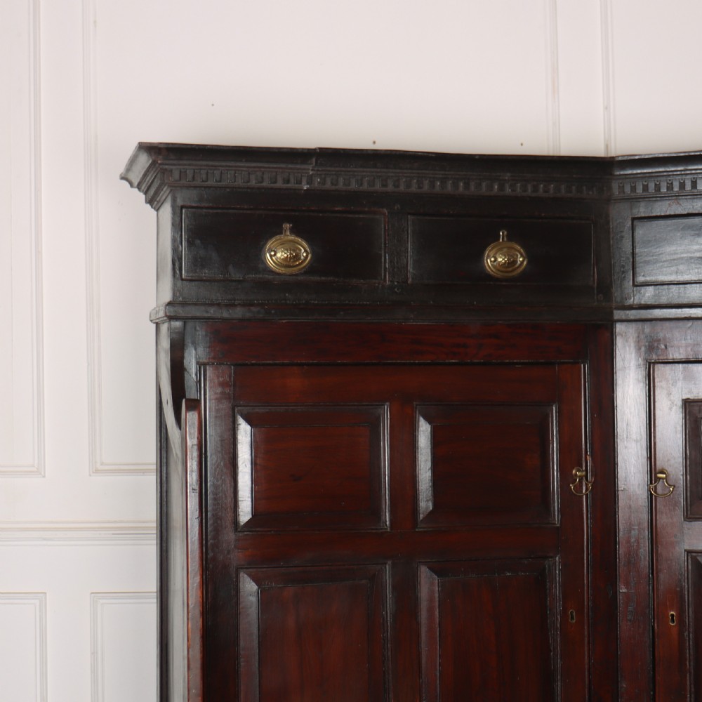 A dark stained oak corner bacon settle featuring high panelled cupboard doors, upper drawers with brass handles, and an integrated bench seat. View III