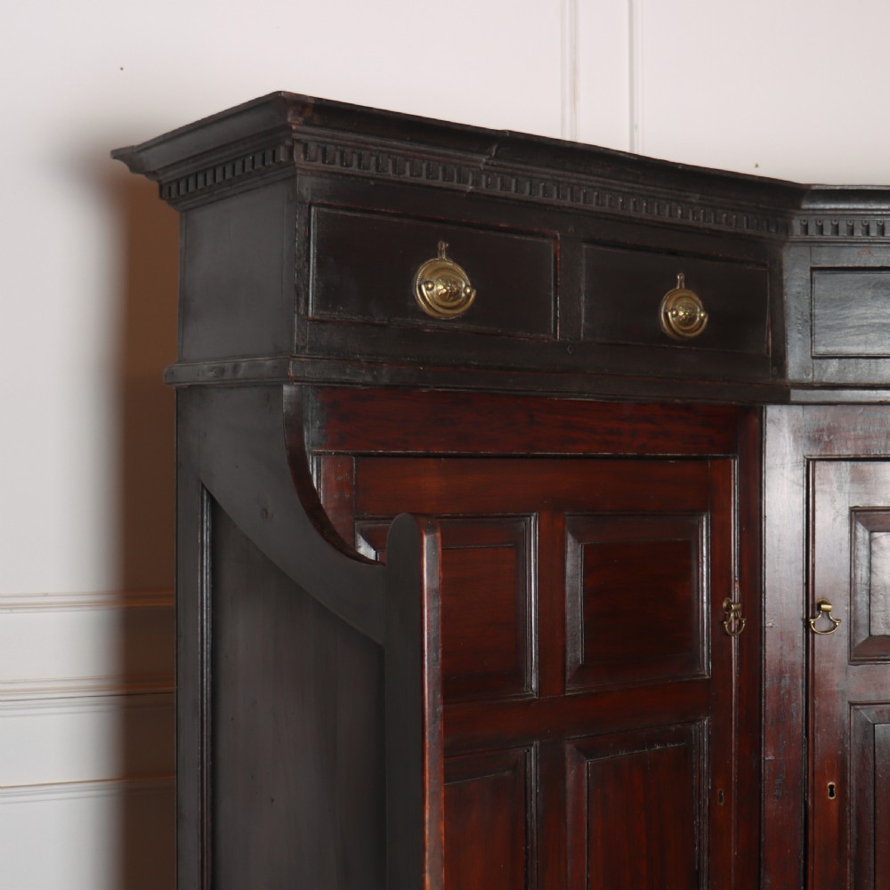 A dark stained oak corner bacon settle featuring high panelled cupboard doors, upper drawers with brass handles, and an integrated bench seat. View V