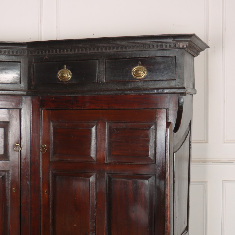 A dark stained oak corner bacon settle featuring high panelled cupboard doors, upper drawers with brass handles, and an integrated bench seat. View VI