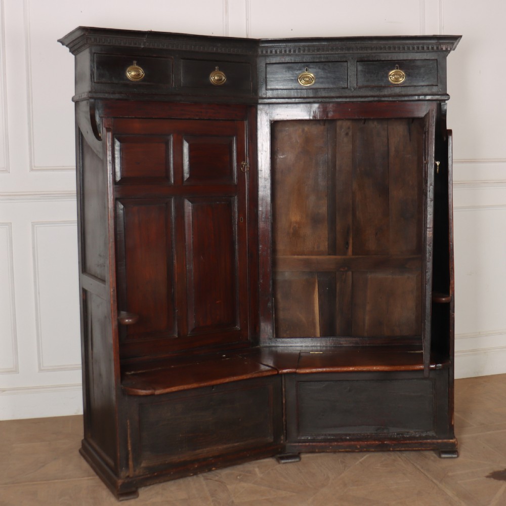A dark stained oak corner bacon settle featuring high panelled cupboard doors, upper drawers with brass handles, and an integrated bench seat. View IX