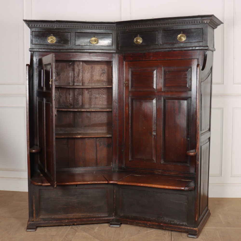 A dark stained oak corner bacon settle featuring high panelled cupboard doors, upper drawers with brass handles, and an integrated bench seat. View X