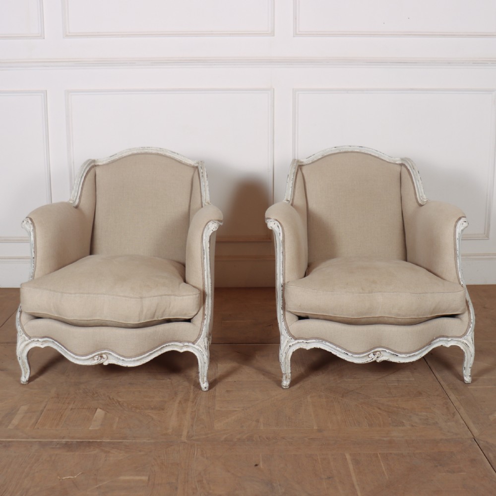 A pair of Swedish Gustavian style armchairs featuring distressed white painted carved wooden frames and beige linen upholstery.