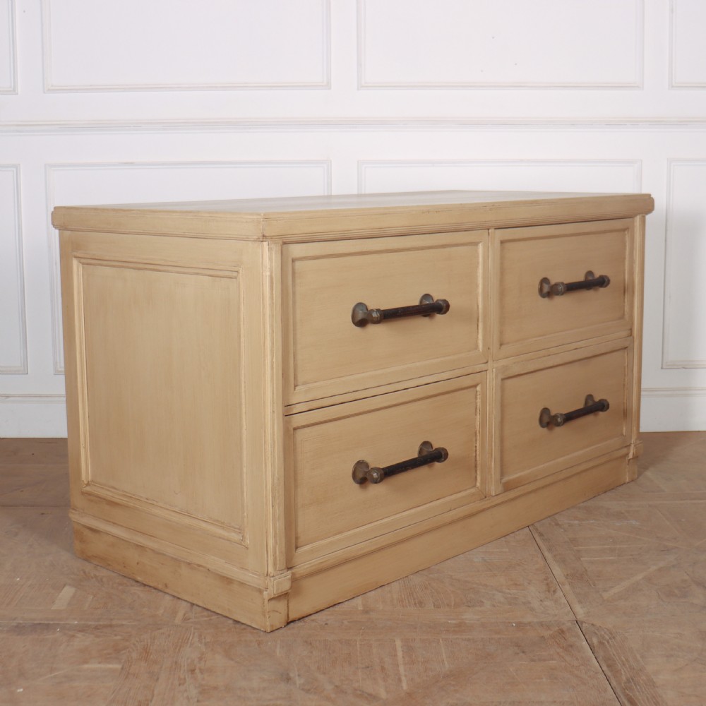A low, wide wooden drapers chest in a neutral beige painted finish featuring four large paneled drawers with dark metal handles.